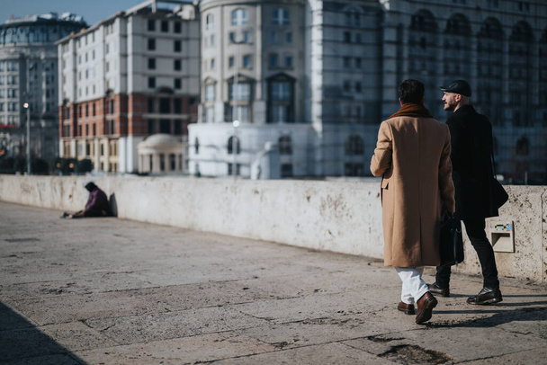 Két divatosan öltözött férfi sétál és beszél egy városi hídon, elmosódott városi épületekkel és egy magányos alakkal a háttérben.. - Fotó, kép