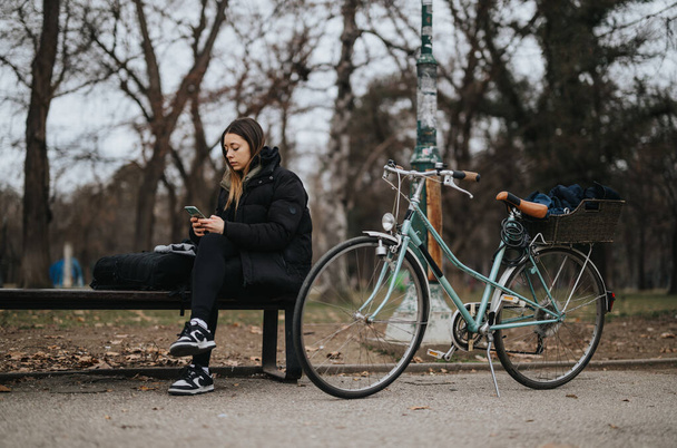 Nuori nainen käyttää älypuhelinta puistossa penkillä vieressä vintage polkupyörän. - Valokuva, kuva