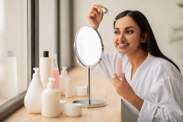 Anti-aging skin care beauty products concept, Attractive long-haired millennial indian woman wearing white bathrobe applying moisturizing serum, nourishing oil on face, looking at mirror at bathroom - Photo, Image