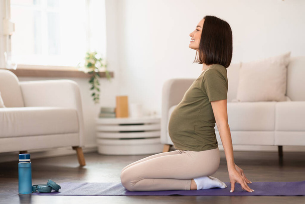 Νεαρή έγκυος γυναίκα εκτελεί μια ανάποδη άσκηση στο στρώμα γυμναστικής στο σπίτι της, με επίκεντρο το τέντωμα και τις πρακτικές αναπνοής, την προετοιμασία του σώματος και του μυαλού για τη μητρότητα. Πλευρική άποψη - Φωτογραφία, εικόνα