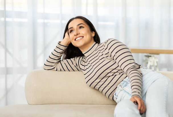 Pensive heureuse jeune femme arabe se trouve sur le canapé dans la lumière salon intérieur, rêve, regarder l'espace libre. Dame profiter de temps libre et de repos, se détendre, pause et style de vie confortable à la maison - Photo, image