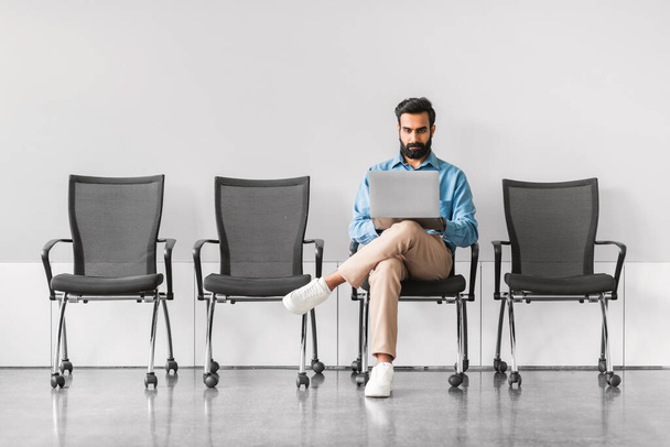 Skupiony indyjski biznesmen korzystający z laptopa siedząc samotnie w przestronnej poczekalni, sugerując przygotowanie do spotkania lub pracy w minimalistycznym otoczeniu - Zdjęcie, obraz