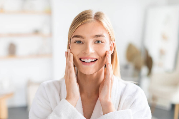Portrait of blonde lady touches cheeks and enjoys her morning skincare routine, beaming with happiness in her home bathroom, smiling to camera. Beauty and pampering concept - Foto, Imagen