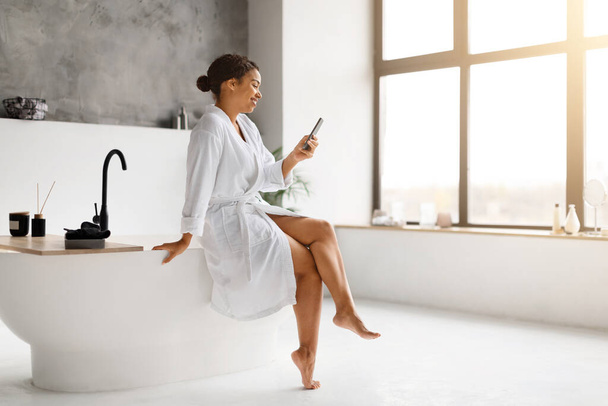 Femme noire en peignoir blanc riant tout en envoyant des SMS sur son smartphone, confortablement assise sur une baignoire moderne dans une salle de bain ensoleillée, message féminin afro-américain heureux avec des amis, espace de copie - Photo, image