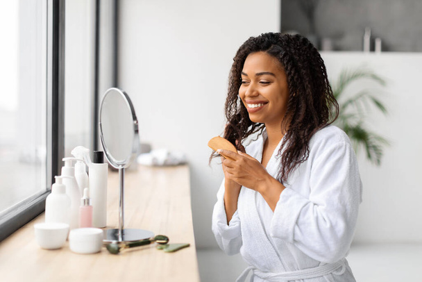 Χαμογελώντας νεαρή μαύρη γυναίκα βούρτσισμα όμορφη σγουρά μαλλιά της με Comb, ενώ κάθεται κοντά στον καθρέφτη στο μπάνιο, Happy African American Female Στο Μπουρνούζι Κάνοντας ρουτίνα περιποίησης μαλλιών στο σπίτι - Φωτογραφία, εικόνα