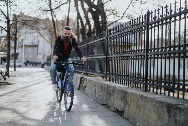 Un homme d'affaires barbu en tenue décontractée roule à bicyclette sur un trottoir ensoleillé de la ville, illustrant les déplacements écologiques. - Photo, image
