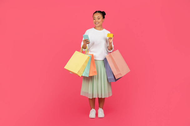 Sorrindo asiático cliente mulher com sacos de compras, smartphone, e cartão de crédito na mão de pé isolado no fundo do estúdio rosa, feliz coreano feminino representando consumidor moderno, comprimento total - Foto, Imagem