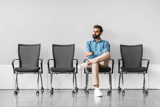 Αποτελείται από Ινδό επιχειρηματία που κάθεται με σταυρωμένα τα πόδια, καθισμένος μόνος του σε μια άδεια σειρά από καρέκλες σε μια αραιή και μοντέρνα αίθουσα αναμονής, πλήρους μήκους - Φωτογραφία, εικόνα