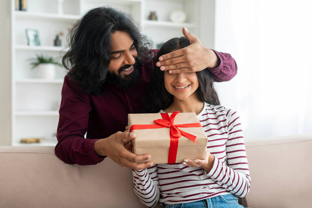 Liebender Millennial-Indianer schließt die Augen seiner Frau, schenkt Geschenkschachtel, macht eine große Überraschung zum Valentinstag, Innenraum, Freiraum. Glücklicher Mann umarmt seine Freundin, hält Geschenk - Foto, Bild