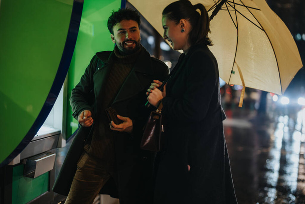 Ein stilvolles Paar an einem regnerischen Tag mit einem Geldautomaten, einer von ihnen hebt Bargeld ab, während er einen Regenschirm hält und ein Gespräch führt. - Foto, Bild