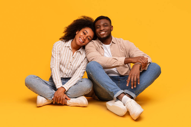 Щаслива афроамериканська пара сидить на схрещених ногах, спираючись один на одного зі змістом посмішок, показуючи випадковий, ласкавий момент на яскраво-жовтому тлі - Фото, зображення