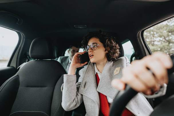 Erkek bir iş arkadaşı arka koltukta oturup, meşgul profesyonelleri resmederken odaklanmış iş kadını araba sürüyor ve telefonla konuşuyor.. - Fotoğraf, Görsel