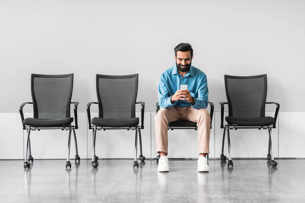 Casual indian businessman seated alone, absorbed in his smartphone, in stark waiting area with rows of empty chairs, suggesting moment of downtime - Photo, Image