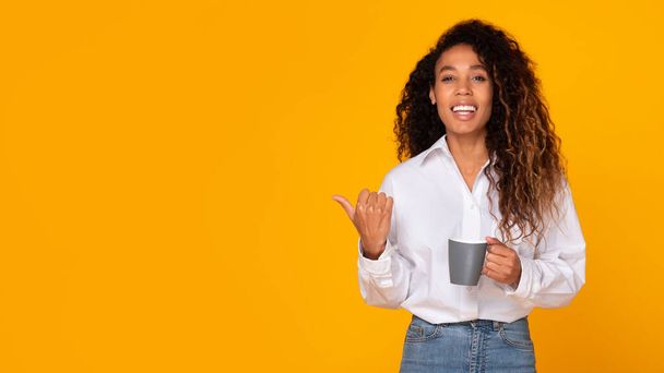 Black Young Lady Holding Coffee Cup Enjoying Hot Drink While Pointing Finger Aside Over Yellow Studio Background, Showing Empty Space For Offer Advertisement. Panorama - Photo, Image