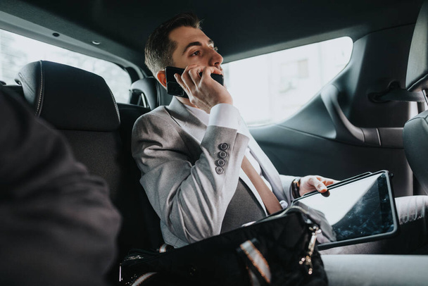 Profesjonalny biznesmen w garniturze korzystający ze smartfona i tabletu podczas podróży samochodem, prezentujący nowoczesny styl życia biznesu. - Zdjęcie, obraz