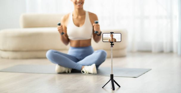 Lächelnde lateinamerikanische Millennial-Frau in Sportbekleidung, die Hanteln hält, während sie ein Fitness-Video mit dem Smartphone auf einem Stativ aufzeichnet und damit moderne Home-Workout-Trends verkörpert, - Foto, Bild