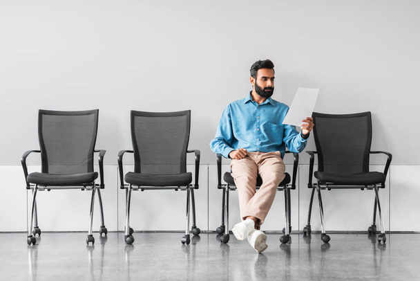 Επαγγελματίας Ινδός με μπλε πουκάμισο κάθεται σε μια άδεια αίθουσα αναμονής, μελετώντας προσεκτικά τα έγγραφα, πιθανόν προετοιμάζοντας μια επερχόμενη συνάντηση ή συνέντευξη - Φωτογραφία, εικόνα