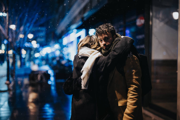 Αγκαλιαστείτε μια χιονισμένη νύχτα της πόλης. Στοργικό ζευγάρι αγκαλιάζει σε εξωτερικούς χώρους. - Φωτογραφία, εικόνα