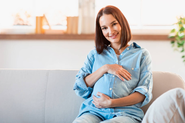 Koncept těhotenství. Usmívající se nastávající matka láskyplně objímající své rostoucí břicho, odrážející radost z nadcházejícího mateřství, pózující obklopená útulnou domácí atmosférou, sedící v obývacím pokoji - Fotografie, Obrázek