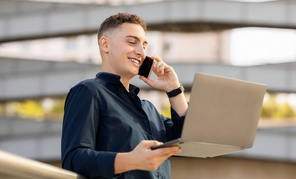 Egy multitasking boldog kaukázusi fiatalember beszél telefonon, miközben egy laptopon dolgozik, kint áll mosolyogva, haditengerészeti inget és okosórát visel. Munka szerkentyű, hívja - Fotó, kép