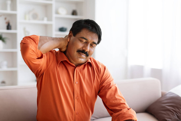 Suffering middle aged indian man feeling neck pain, sitting on couch in cozy living room at home. Chronic injury, muscle inflammation strain and backache concept, copy space - Photo, Image