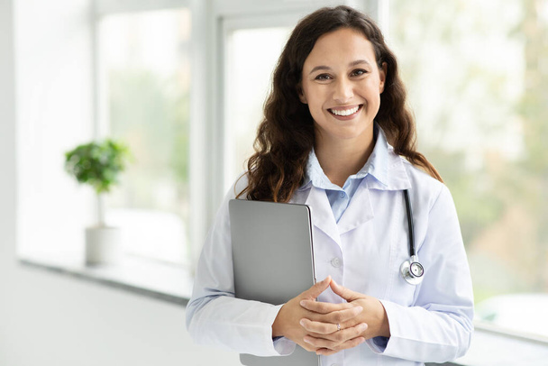 Moderne technologieën in de gezondheidszorg. Glimlachende jonge vrouwelijke arts met laptop poserend in haar kantoor door het raam, kopieerruimte. Jonge vrouw therapeut holding computer, kliniek interieur - Foto, afbeelding