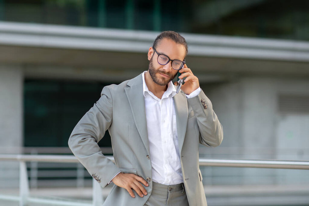 Komoly öltönyös, fiatal üzletember, aki a szabadban telefonál, magabiztos férfi vállalkozó, aki csípőn tartja a kezét, városi háttérrel szemben áll, aktív üzleti kommunikációt mutat be - Fotó, kép