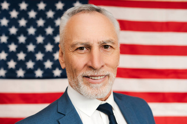 Egy jóképű, idős amerikai férfi portréja, magabiztos politikus, aki amerikai zászlóval a háttérben nézi a kamerát. A választás napjának koncepciója  - Fotó, kép