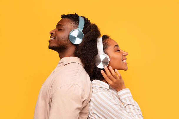 Relajada joven pareja negra de pie espalda con espalda, cada uno con auriculares, perdido en la música con sonrisas de contenido en un fondo amarillo soleado, vista lateral - Foto, imagen