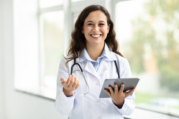 Technologies modernes dans l'industrie de la santé concept. Jeune femme médecin souriante avec tablette numérique posant dans son bureau par fenêtre, espace de copie. Jeune femme thérapeute tenant tampon numérique, intérieur de la clinique - Photo, image