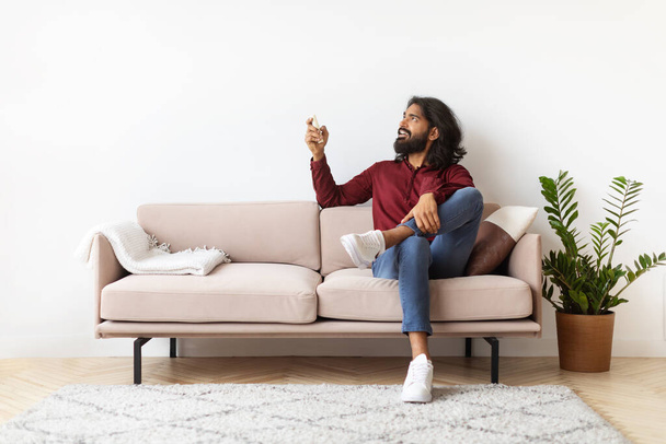 Uvolněný tisíciletý vousatý dlouhosrstý indián sedící na gauči v útulném interiéru obývacího pokoje, zapínající klimatizace s dálkovým ovládáním, zaměřující se na kopírování prostoru na prázdné bílé stěně, chlap chladící doma - Fotografie, Obrázek