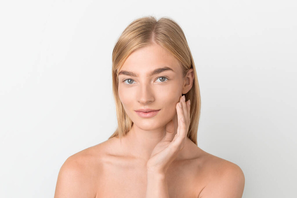 Skincare Concept. Beautiful Blonde Woman With Smooth Skin Touching Face With One Hand, Enjoying Softness Of Cheek, Posing Shirtless With Natural Makeup Over White Studio Background - Photo, Image