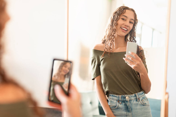 Genç kız akıllı telefonuyla mutlu bir şekilde aynanın yanında poz vererek selfie çekiyor, sosyal medya için gençlik yaşam tarzını içeride yakalıyor. Bloglar ve aletler konsepti. Seçici odak - Fotoğraf, Görsel