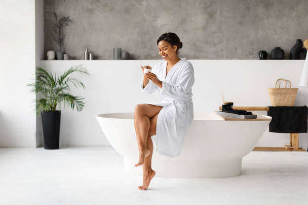 Pielęgnacja skóry. Szczęśliwa młoda Afroamerykanka stosująca krem w łazience, uśmiechnięta czarna kobieta nawilżająca skórę po kąpieli, siedząca na luksusowej wannie, mająca dzień piękna w domu, przestrzeń do kopiowania - Zdjęcie, obraz