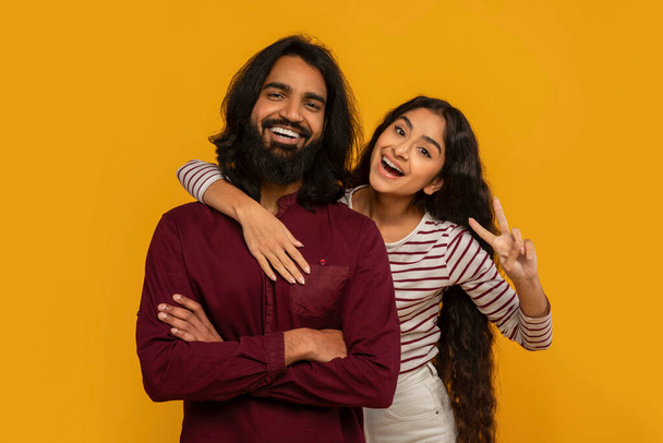Portrait of cheerful young indian man and woman lovers embracing on yellow studio background, smiling at camera, gesturing showing peace V gesture. Love, relationships, affection - Photo, Image