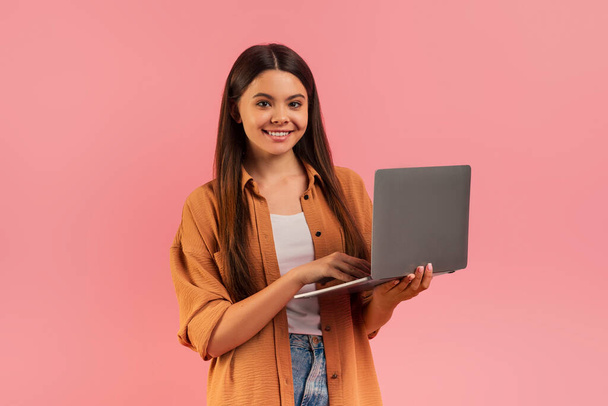Pewna siebie nastolatka trzymająca laptopa i uśmiechnięta do kamery, piękna nastolatka nosząca luźny strój stojąca na różowym tle studia, reprezentująca edukację online lub pracę zdalną - Zdjęcie, obraz