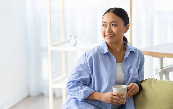 Happy beautiful millennial korean woman relaxing at home, sitting on couch with mug in her hands in cozy living room, drinking coffee, looking at copy space for advertisement - 写真・画像