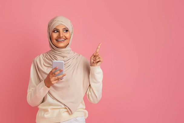 Innoissaan musliminainen Hijab Holding älypuhelin ja osoittaa Kopioi Space, Hymyilevä arabia Lady nostamalla sormi ylös, ottaa idea, Nauttii Mobile Tarjous, Seisoo Pink Studio Tausta - Valokuva, kuva