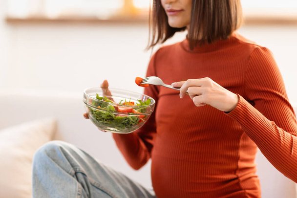 Nierozpoznana ciężarna kobieta jedząca świeżą sałatkę wegetariańską z miski, siedząca na kanapie w salonie w domu, przycięta. Oczekująca Pani ciesząca się zdrowym odżywianiem w czasie ciąży - Zdjęcie, obraz