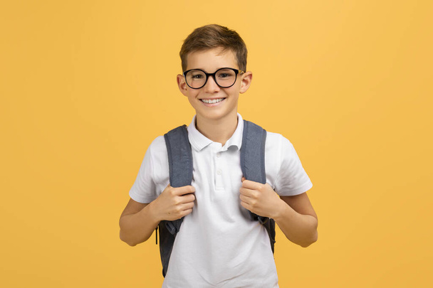 Alegre colegial con gafas y mochila de pie con confianza en el fondo del estudio amarillo, niño adolescente feliz listo para la escuela, disfrutando del aprendizaje y la educación, espacio para copiar - Foto, Imagen