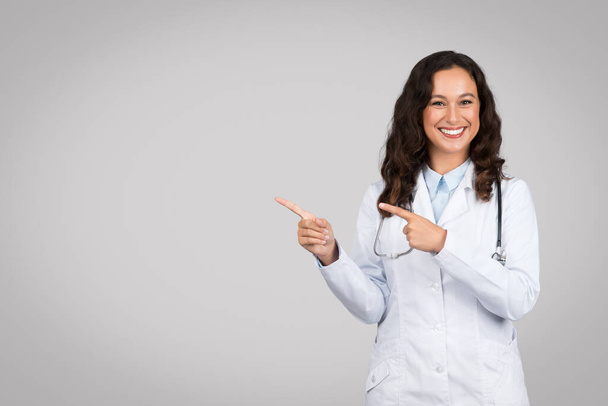 Medico donna radiante in camice bianco che punta gioiosamente al lato dello spazio libero con entrambe le mani, stetoscopio intorno al collo su uno sfondo grigio - Foto, immagini