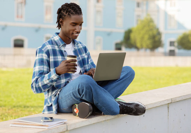 Glücklicher schwarzer Student, der online mit Laptop-Computer lernt, sich auf Prüfungen im Freien vorbereitet, auf Brüstung mit Papierkaffeetasse sitzt, während er in der Nähe des Universitätscampus im Internet surft - Foto, Bild