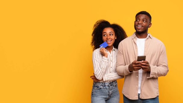 Захоплена афроамериканська пара тримає кредитну картку і смартфон, посміхаючись і дивлячись на вільний простір, розмірковуючи про покупку в Інтернеті на яскраво-жовтому тлі - Фото, зображення