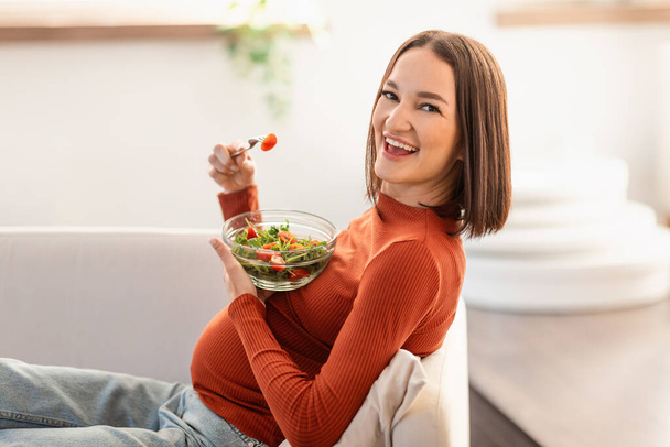 Ravitsemus raskauden aikana. Onnellinen nuori raskaana oleva nainen syö kasvissalaattia istuessaan sohvalla kotona, nauttien terveellisestä orgaanisesta vitamiiniruoasta ja rentoutumisesta olohuoneessa - Valokuva, kuva