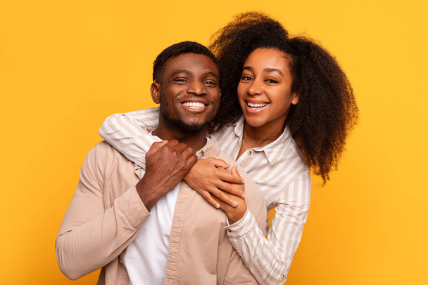 Χαρούμενο νεαρό μαύρο ζευγάρι που μοιράζεται μια αγκαλιά, με φωτεινά χαμόγελα και στενό δεσμό, σε ένα ζωντανό κίτρινο φόντο - Φωτογραφία, εικόνα