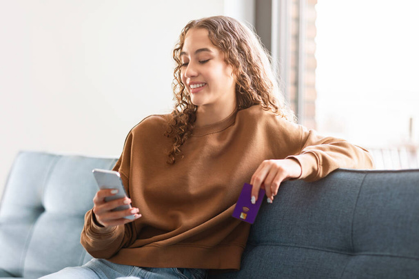 Valkoihoinen teini tyttö ostoksia verkossa matkapuhelimella tilalla luottokortti, tehdä ostoksia, lounging sohvalla kotona, käsittää helppous modernin verkkokaupan ja digitaalisen kulutuksen - Valokuva, kuva