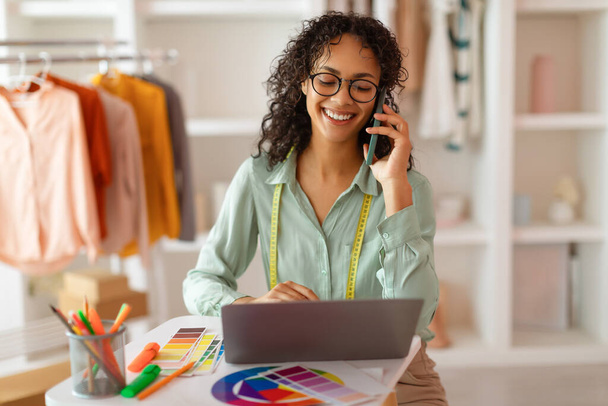 Designer de moda engajada em óculos conversando no telefone enquanto trabalhava em seu laptop, cercada por amostras de cores vibrantes em seu estúdio - Foto, Imagem