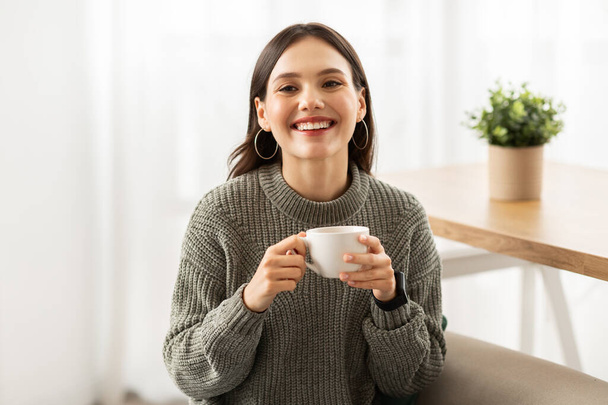 Portrait de joyeux jolie jeune femme européenne assise sur le canapé à côté de la fenêtre tenant tasse de café et souriant à la caméra, boire du thé chaud, intérieur de la maison, espace de copie - Photo, image