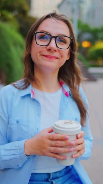 El retrato de la mujer en gafas sostiene el café para ir en las manos y mirar a la cámara. Vídeo vertical. Movimiento lento - Metraje, vídeo