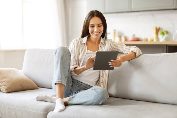 Tartalom fiatal nő böngészés internet digitális tabletta, ül keresztbe lábú kényelmes kanapén kényelmes nappali belső, boldog Y-generációs női pihentető modern modul otthon, másolás tér - Fotó, kép